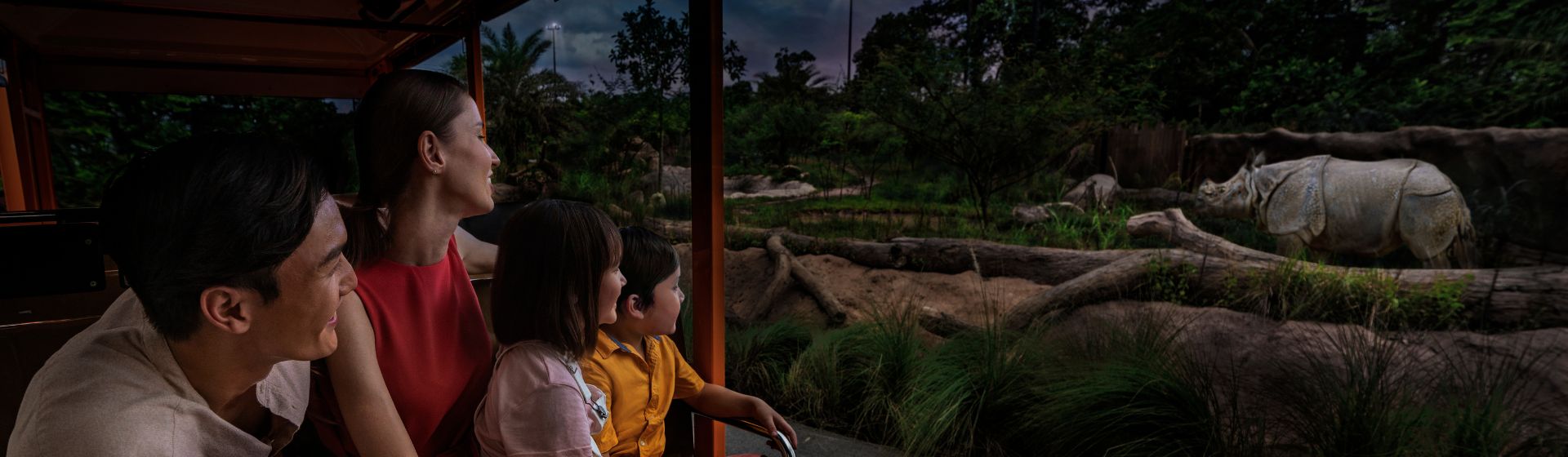 A family of 2 parents with 2 young kids riding a tram past the indian rhino enclosure at at Night Safari in Singapore